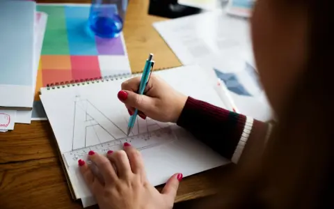 Ilustrator pracuje nad projektem, zamieniając litery na krzywe. Na stole leżą przybory do rysowania i próbnik kolorów.