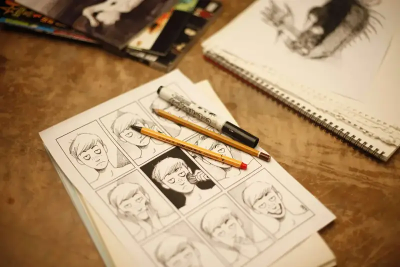 Panel komiksowy z różnymi wyrazami twarzy, narysowany jedną kreską. Obok leżą ołówki i marker.